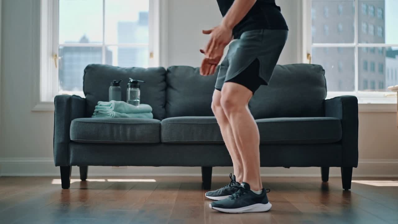 Uomo che inizia uno squat basso in abbigliamento sportivo mentre fa squat a casa per il fitness con divano e bottiglie d'acqua