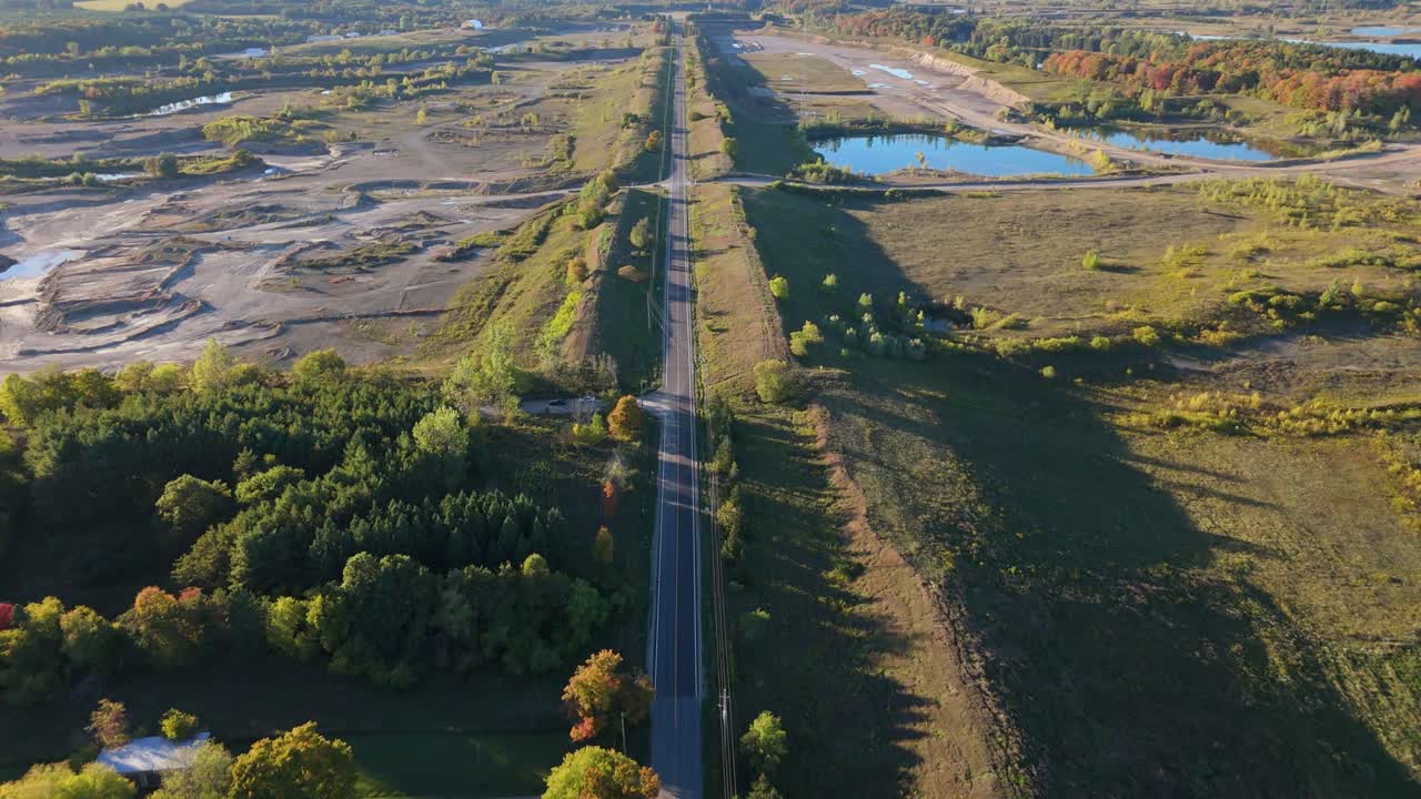A stunning aerial view of a long road in Caledon, Ontario, surrounded by vibrant fall colours during a golden hour sunset. The road separates a gravel pit from the natural landscape