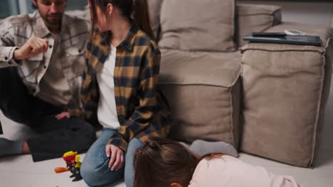 una pequeña chica morena en un traje rosa dibuja en el suelo en papel mientras sus padres se comunican entre sí y se sientan cerca de la chica cerca del gris-marrón en un apartamento estudio moderno