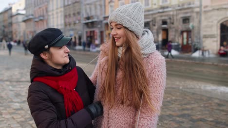 Two-smiling-women-tourists-walking-together-on-city-street,-family-couple-talking,-embracing