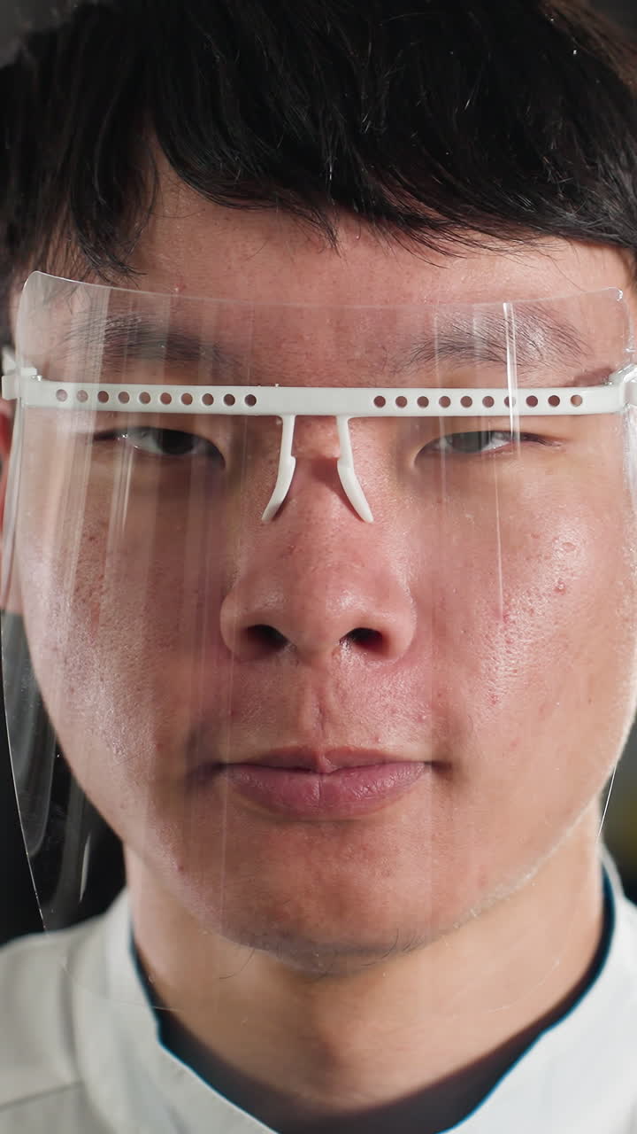 색 실험실 코트와 보호 얼굴 방패를 입은 실험실 기술자의 클로즈업, 대담한 표정으로 박이는, 흐릿한 배경과 함께 산업 환경, 부드러운 조명