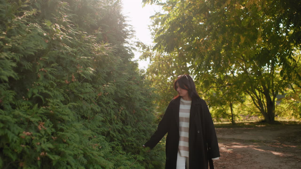 겨울 코트를 입은 차분한 여성이 공원 산책로를 따라 걷고 있어요. 손은 푸른 울타리 옆 가을 단풍 위를 스치듯 지나가고, 햇살이 나뭇잎 사이로 부드럽게 비쳐 따뜻한 햇살 아래에서 야외 산책을 즐기는 모습이에요.