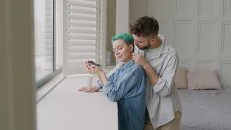 young man tenderly cuddling his girlfriend who holding a mug while standing together next to the window in the bedroom