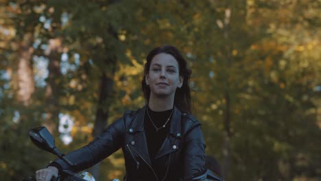 Pretty-smiling-European-young-woman-driving-a-motorbike-wearing-leather-jacket-in-forest-with-vibrant,-colorful-golden-autumn-leaves-on-sunny-day