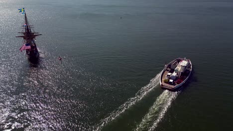 An-aerial-view-of-a-17th-century-wooden-ship-and-a-Greenport-North-Ferry,-both-heading-out-on-Greenport-Harbor-on-Long-Island-on-a-sunny-day