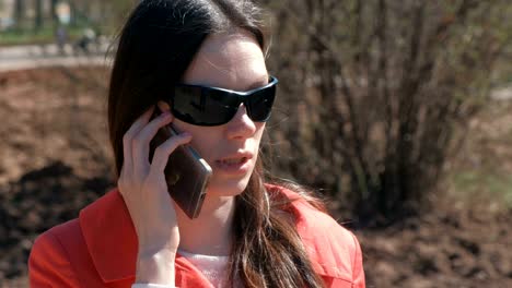 retrato de una joven morena con gafas de sol hablando por teléfono en el parque de primavera.