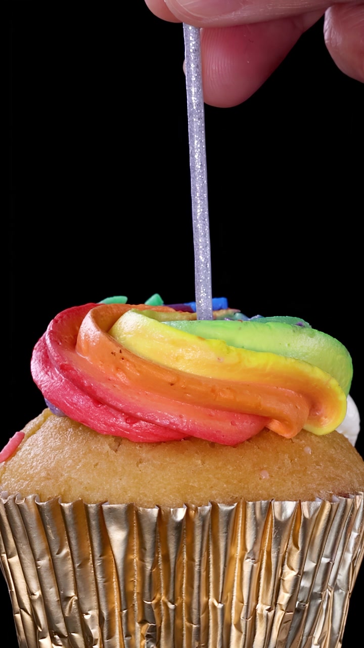 Una mano inserisce una candela argentata in un cupcake con glassa arcobaleno e pirottino dorato, ripreso in primo piano su sfondo nero con illuminazione uniforme