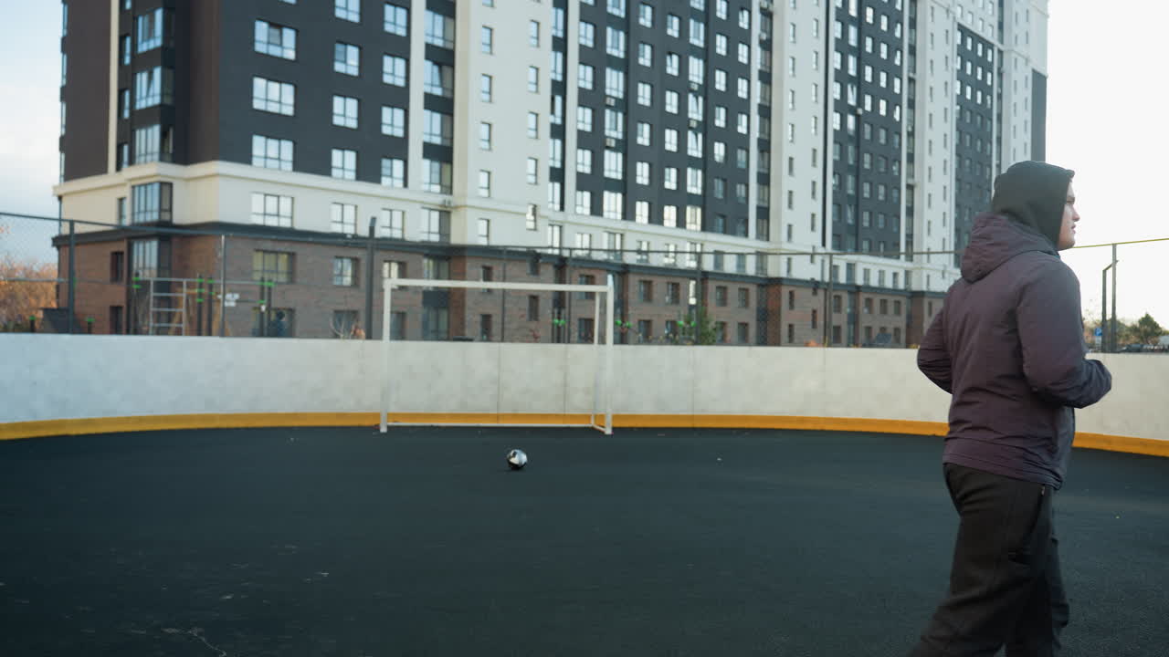 спортсмен бегает на открытой арене с футбольным мячом и головным столбом в фокусе, окруженный городскими многоэтажными зданиями, освещением, спортивной деятельностью и городским образом жизни в оживленной среде