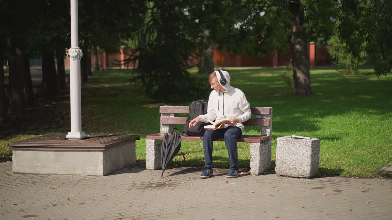 공원 벤치에 앉아 있는 백인 남성 직장인이 백팩에서 우산을 꺼내 펼치고, 빗방울이 떨어지기 시작하자 휴대폰을 확인하고 있습니다. 헤드폰을 착용하고 있으며, 가로등 기둥과 나뭇가지 캐노피가 머리 위에 드리워져 있습니다. 캐주얼 재킷과 청바지 차림입니다.