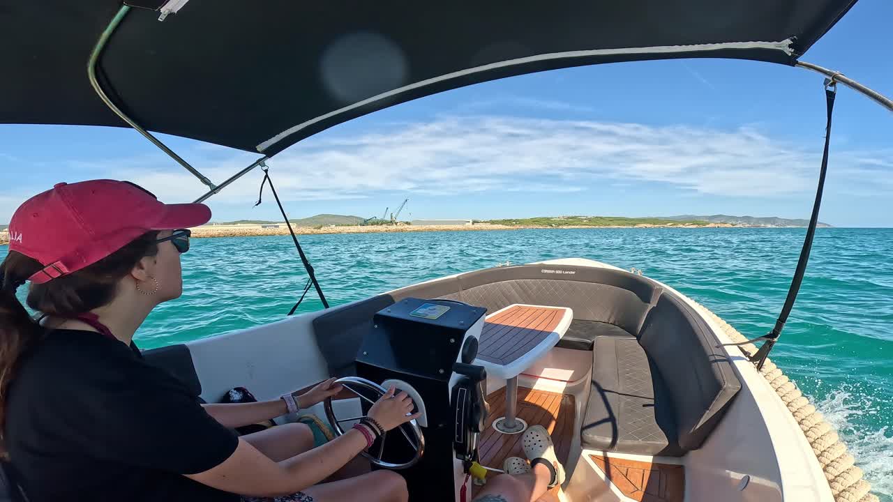 Young woman in red cap and sunglasses steers a boat on a sunny day, gliding over calm waters with clear blue sky and scenic coastline in the background