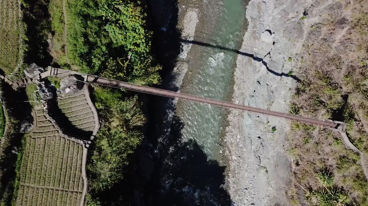 Top down following metal suspension bridge spanning over mountain river creek leading to trails farms rice patties vegetable gardens trails on sunny day