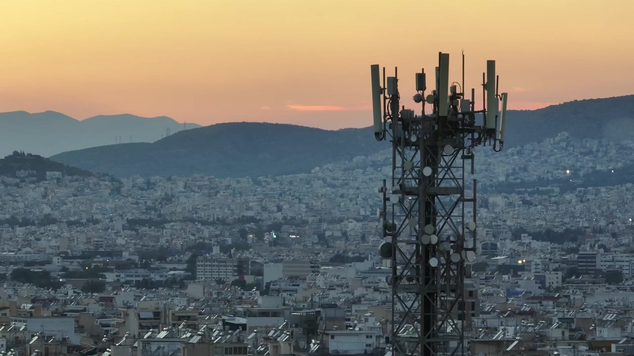 Drone footage circling communication antennas at sunset, showcasing the vibrant Athens cityscape, golden sky, and panoramic urban landscape from above.