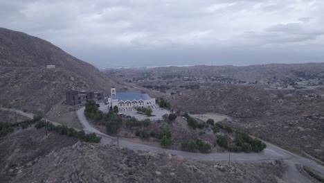 a drone gracefully pulls away from a church atop a hill on a cloudy day, capturing the serene beauty of the hillside setting