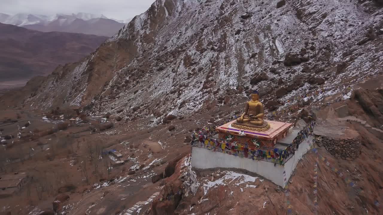 Gyllen Buddha-statue plassert på en steinete, snødekt klippe ved Hemis-klosteret i Ladakh på en vindfull vinterdag, omgitt av snødekte fjell, fargerik baseplattform og bønneflagg