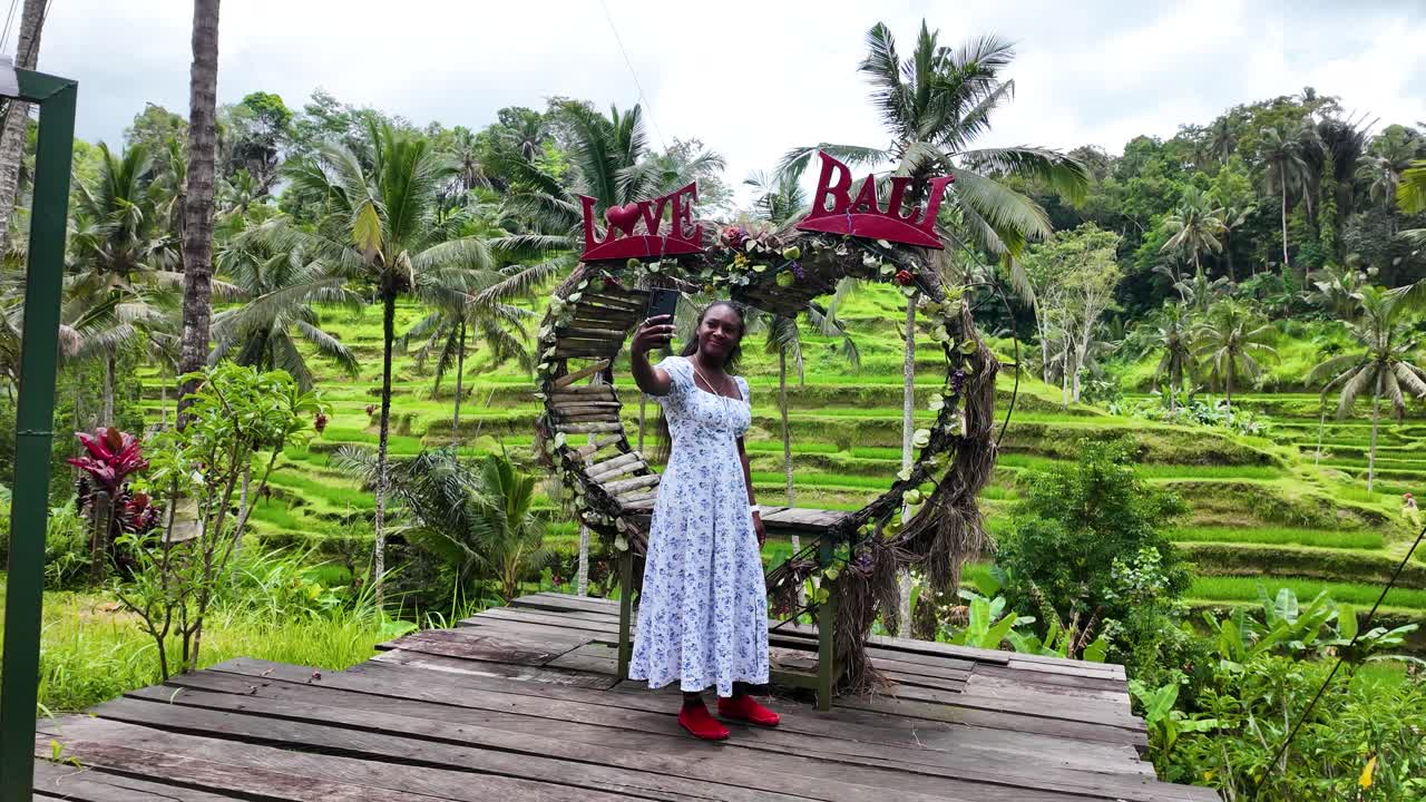 Woman takes selfie at Bali's Tegallalang Rice Terrace with palm trees under a wooden heart