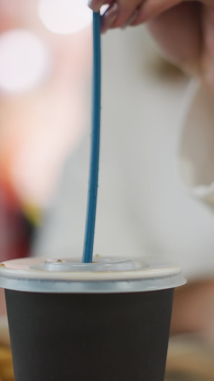 Close-up of hand delicately inserting a blue straw into a takeaway tea cup lid, set against a softly blurred background with vibrant bokeh lights