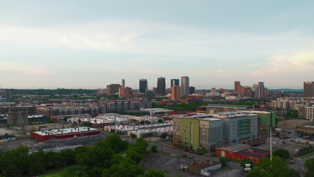 Flygfoto över Birminghams, Alabamas stadssiluett i mjukt kvällsljus – centrumtorn reser sig bortom nya lägenheter, lagerbyggnader och lummiga bostadsområden.