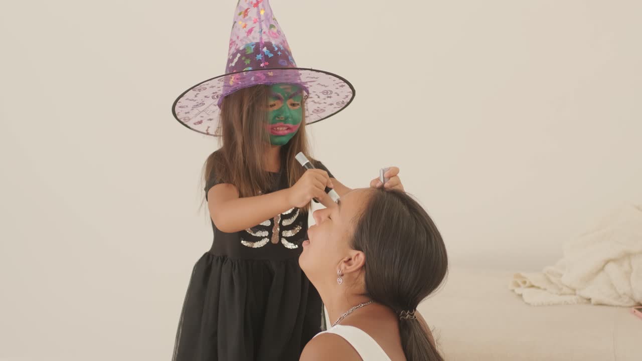 niña con sombrero de bruja aplicando maquillaje a su madre embarazada para halloween