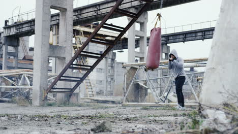 distant view of caucasian man in grey hoodie hitting a punching bag outdoors an abandoned factory on a cloudy morning