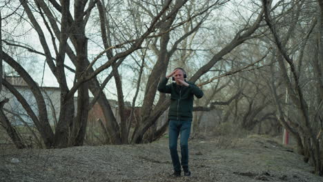 a man in a green jacket with headphones dances energetically in a wooded area, the ground is covered with leaves and twigs