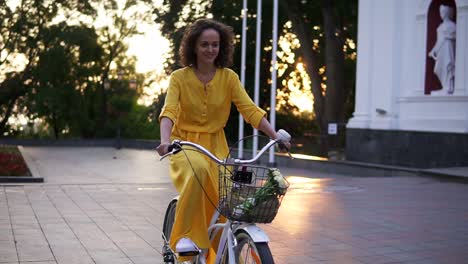 joven mujer atractiva está montando una bicicleta de la ciudad con una canasta y flores en el parque de la ciudad mientras el hijo está levantando. destello de la lente