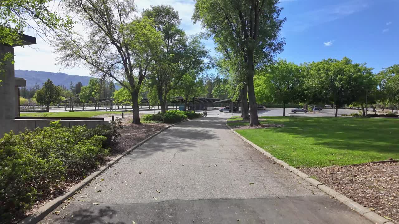 Perspectiva em primeira pessoa caminhando por um caminho pavimentado através do campus verde e ensolarado do West Valley College em Saratoga, Califórnia, em um belo dia de primavera com árvores e um céu azul.