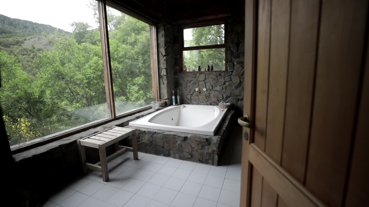 Opening door and entering luxury bathroom with bathtub fitted along window wall facing beautiful dense greenery