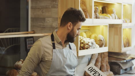 Männlicher-Junger-Bäckereiverkäufer,-Der-Mit-Einem-Schild-Aufwirft,-Das-Innerhalb-Des-Brotladens-Geöffnet-Ist