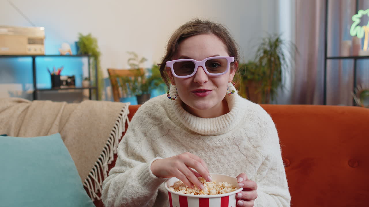 Woman sitting on couch eating popcorn and watching interesting tv 3d film sport game online at home