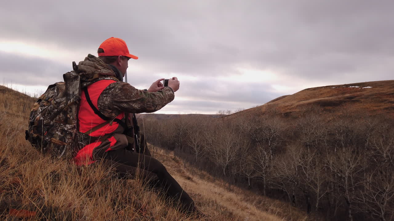 Young Caucasian male hunter with rifle wearing neon orange hat and vest sitting in field taking photos with mobile handheld phone on cloudy day, Saskatchewan, Canada, static close up