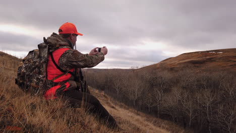 young caucasian male hunter with rifle wearing neon orange hat and vest sitting in field taking photos with mobile handheld phone on cloudy day, saskatchewan, canada, static close up