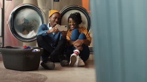 young couple sitting on floor with basket of dirty clothes and watching video on smartphone while washing machines working man and woman using phone and resting in lundry service