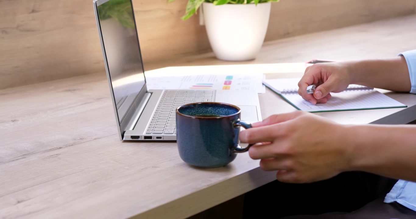 Asian man in light-blue shirt adjusting pen then reaching for blue mug to drink at desk