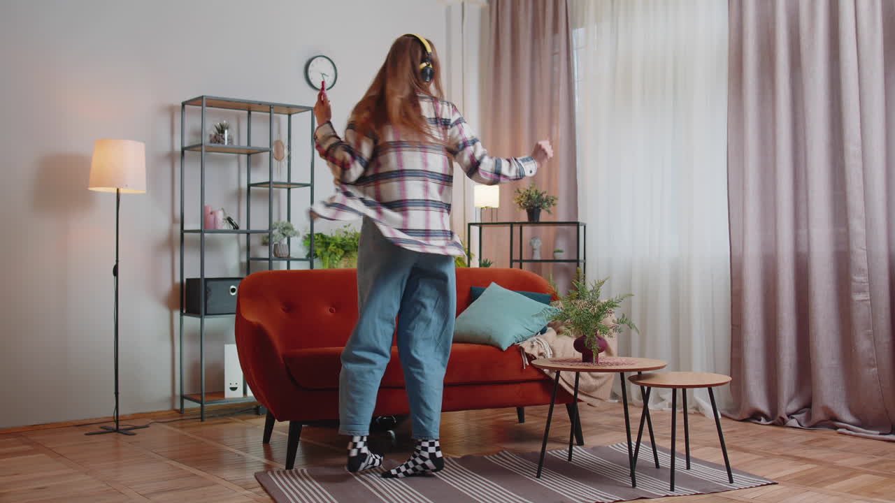 Overlykkelig ung kvinde med trådløse hovedtelefoner danser og synger på en hyggelig sofa i stuen derhjemme.