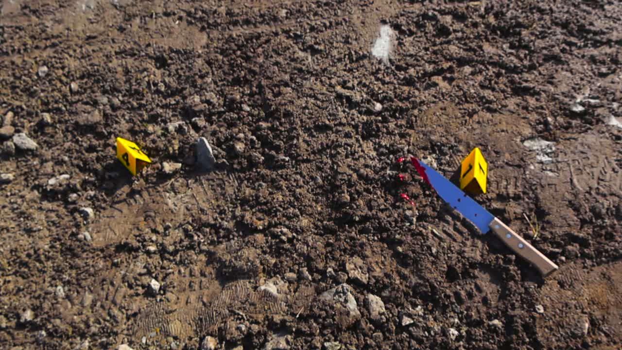Top down view of a bloody knife and a footprint next to police yellow colored crime markers on muddy dirt covered ground. Sunny day in a rural countryside area after a homicide has taken place