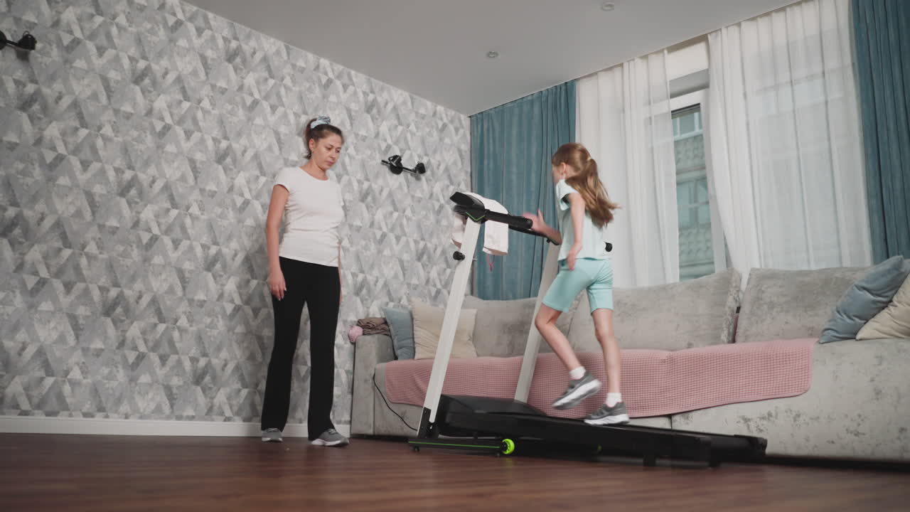 Kid jogging on treadmill set beside cushioned sofa while woman coach stands nearby offering guidance during indoor workout session in well-lit living room with patterned wall and large window