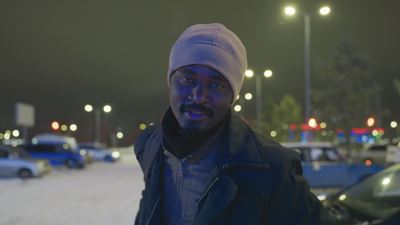 Young man wearing winter hat and jacket outdoors at night adjusts bag strap while smiling, illuminated by city streetlights and blurred background with cars and snow