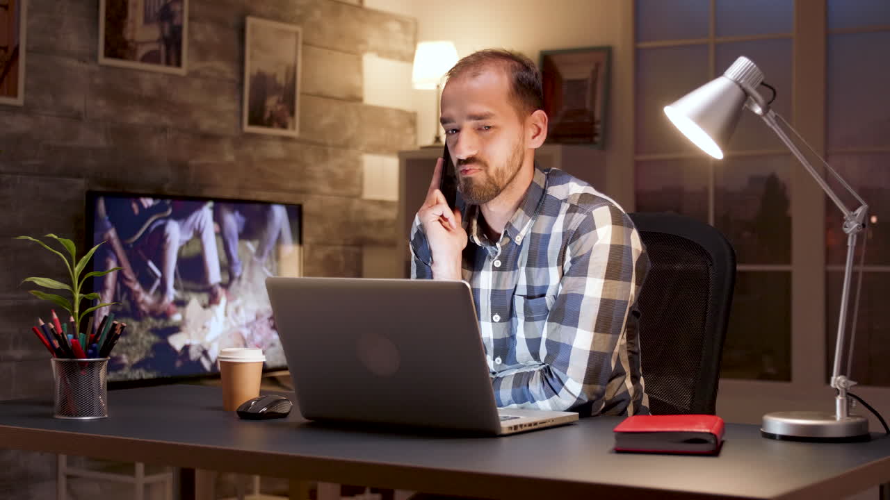 Mand, der arbejder ved et skrivebord på sin bærbar computer, mens han taler i telefon om natten
