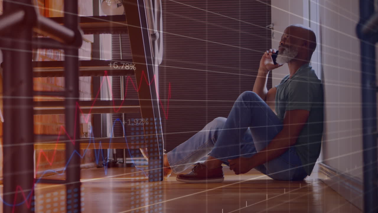 Animation of graphs, numbers, senior african american man sitting on floor and talking on cellphone