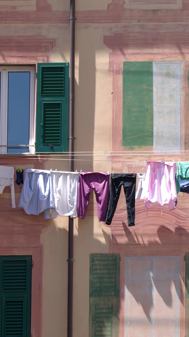 Static medium shot of colorful laundry drying outside a pink facade with green shutters.