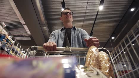 Caucasian-young-guy-in-glasses-and-stylish-blue-shirt-walks-by-the-store-with-shopping-trolley.-Passing-through-the-goods-aisle-of-a-big-supermarket.-Low-angle-footage-from-the-moving-shopping-cart