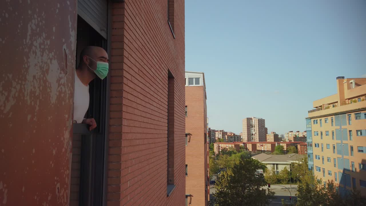 A lateral shot of a man leaning out the window in a flat, taking off a mask and breathing deeply