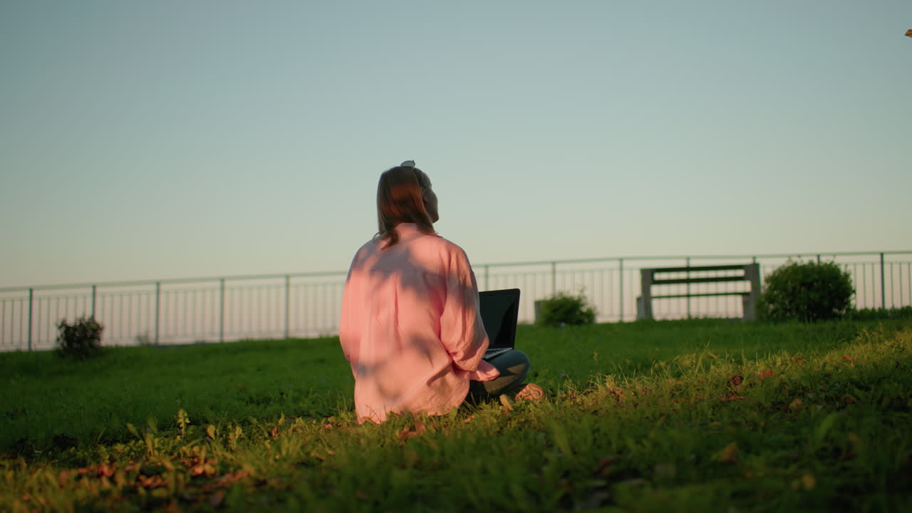 핑크 을 입은 프리랜서와 안경을 머리에 고 노트북을 다시 쳐다보고, 옷에 그림자를 던지는 빛, 먼 배경의 벤치와 철 레일링의 뒷면