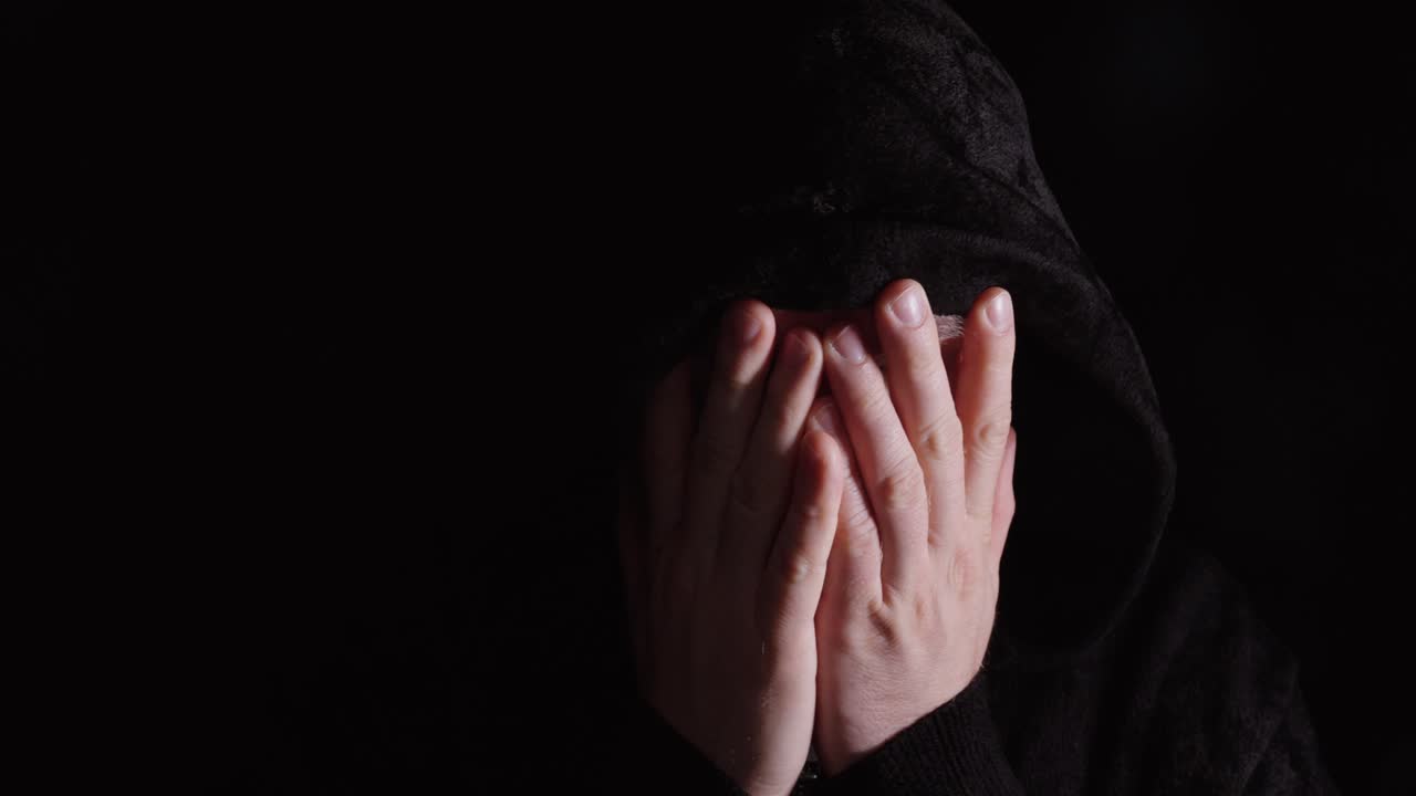 Stemningsfull saktefilm av en mann i mørk hettegenser som ser ned og rister på hodet, uttrykker tristhet, skuffelse eller anger. Ideell for emosjonelle, psykisk helse- og dramatiske konsepter