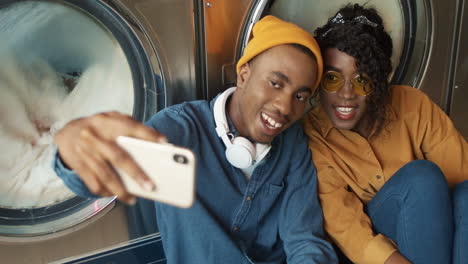 Pareja-Alegre-Sonriendo-A-La-Cámara-Del-Teléfono-Inteligente-Mientras-Toma-Una-Foto-Selfie-En-El-Servicio-De-Lavandería