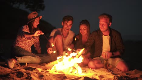 jóvenes amigos alegres sentados junto al fuego en la playa por la noche, cocinando malvaviscos en palos juntos. filmado en 4k