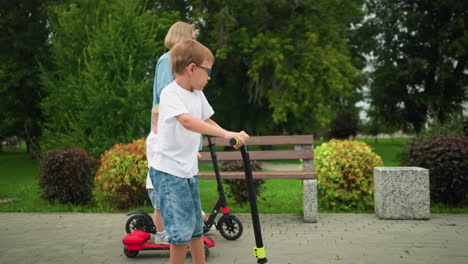 a mother and her children ride scooters along a park path, with the younger child working hard to keep up