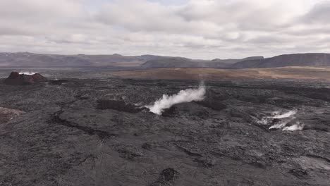 aerial view of black lava field in iceland with white smoke rising from cracks, volcanic terrain, cloudy sky, raw nature