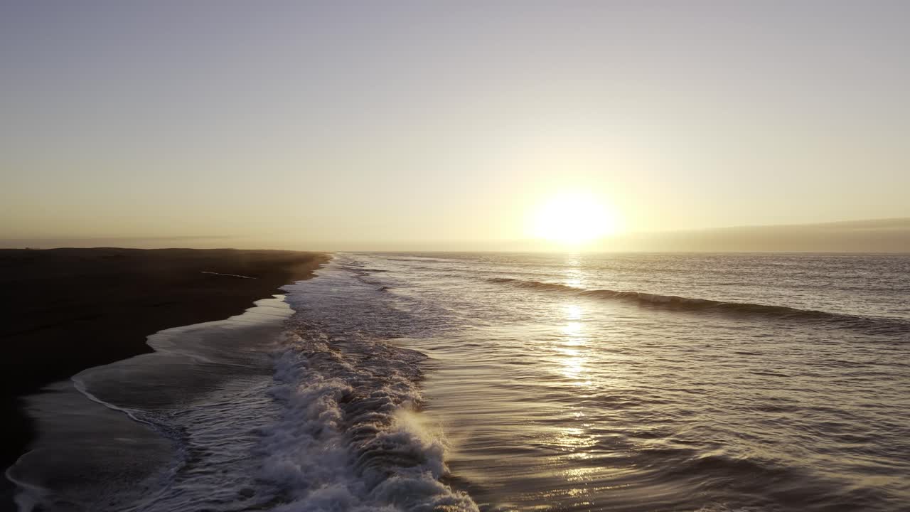 Deslumbrante tomada de drone em baixa altitude voando ao longo de uma praia de areia escura perto das Dunas de Putú, Maule. Ondas quebrando sob um pôr do sol brilhante. Chile, 4K