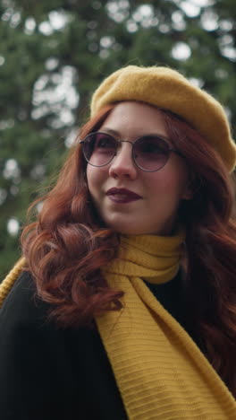 dama elegante con el cabello rojo largo y ondulado, con un abrigo negro y boina amarilla con pañuelo a juego, toca su pañuelo mientras mira hacia arriba, caminando a través de un parque vibrante adornado con colorido follaje de otoño